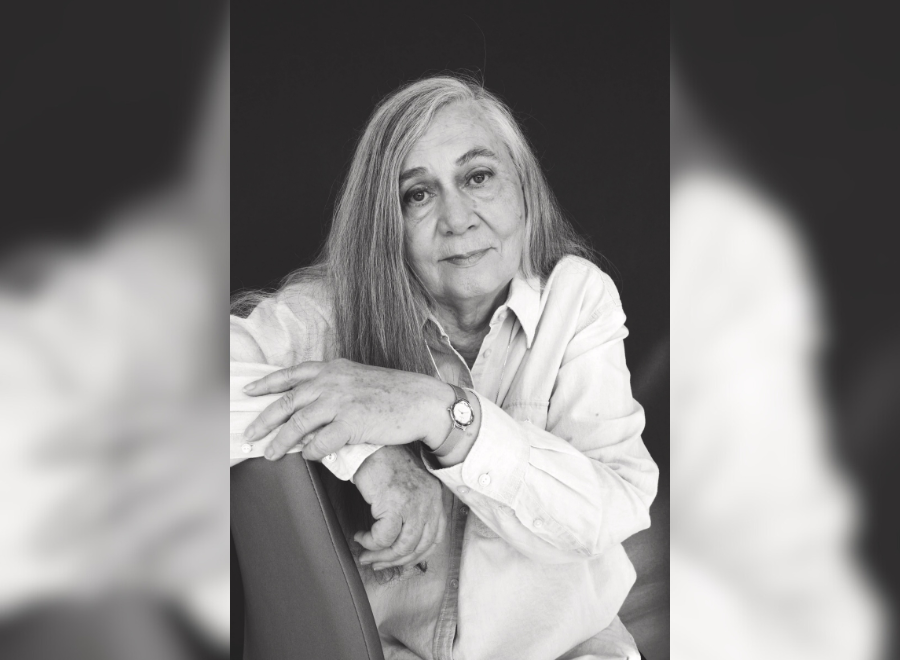 A middle-aged white woman with grey hair poses on a chair. The photo is in black and white. She is wearing a light-coloured button up and a watch on her left hand. Her left hand rests on her right arm.
