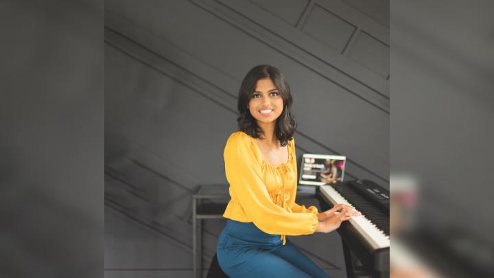 A young brown-skinned woman sits in front of a piano. She is wearing a yellow long-sleeved top with blue pants. She is smiling and has shoulder-length dark brown hair. Her hands are on the piano keys.