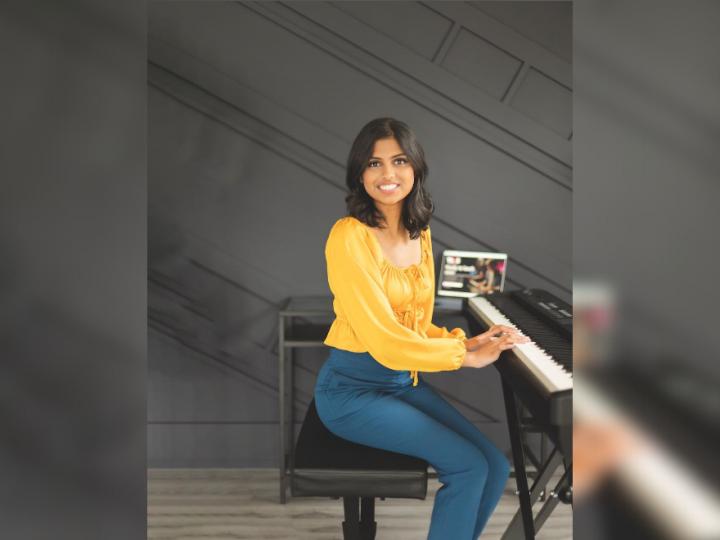 A young brown-skinned woman sits in front of a piano. She is wearing a yellow long-sleeved top with blue pants. She is smiling and has shoulder-length dark brown hair. Her hands are on the piano keys.