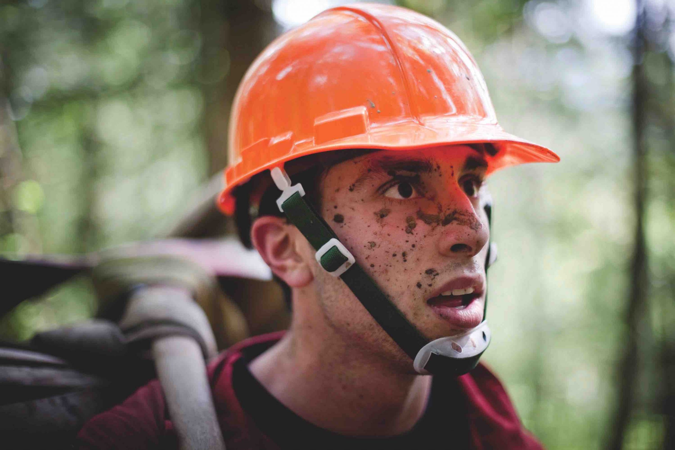 Close up of a young man with an orange helmet and mud splattered on his face. He is carrying a large backpack and surrounded by trees. 