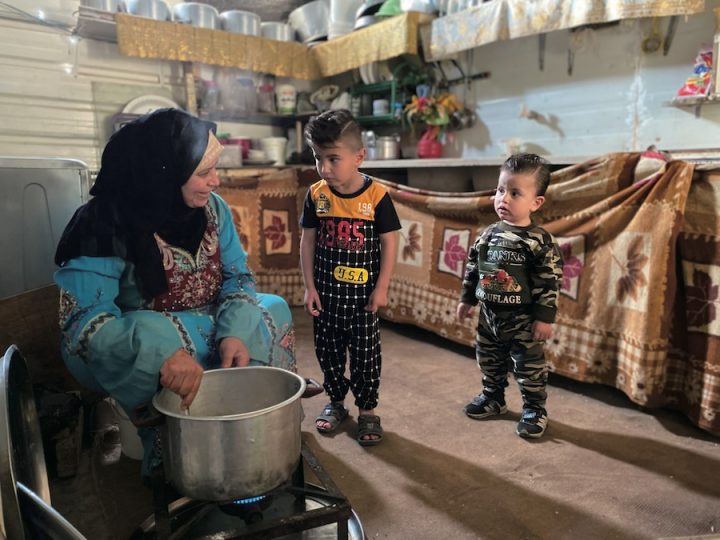 A brown-skinned woman in a navy headscarf and blue dress sits in front of a pot over a gas stove. She is holding a spoon and stirring the contents. She is smiling and talking to two little boys. One boy is older and taller. He is dressed in a matching shirt and sweatpants with black sandals on. The other boy is smaller and younger. He is wearing a camouflage sweatshirt with matching sweatpants and blue sneakers.