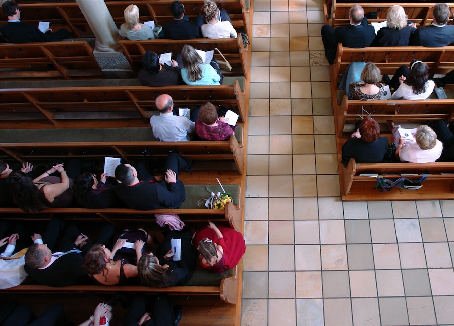 Aerial view of a church congregation. The floor has different coloured tiles, you can see the church pews are filled with people and the tops of their heads.