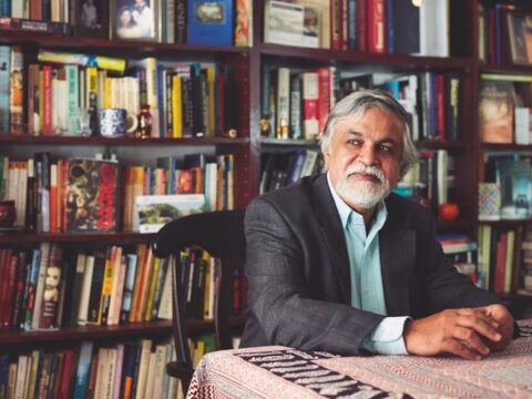 A man sits in front of a bookshelf wearing a blue collared shirt and a blazer. His hands are folded on the table in front of him.