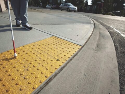 Low perspective of a sidewalk showing yellow tactile bumps, a white cane for a visually-impaired user. You can see a person's legs and black sneakers holding the cane.