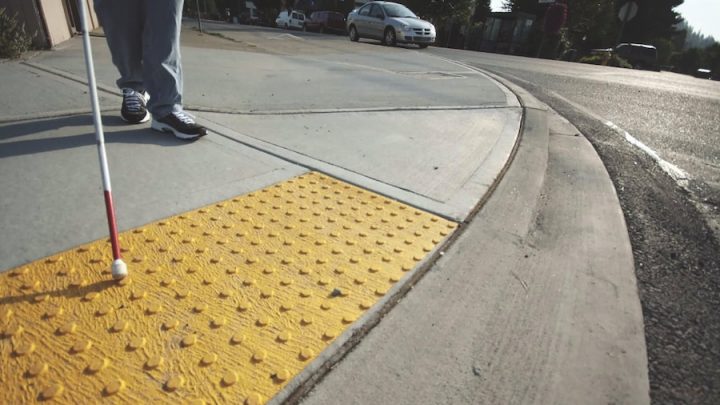 Low perspective of a sidewalk showing yellow tactile bumps, a white cane for a visually-impaired user. You can see a person's legs and black sneakers holding the cane.