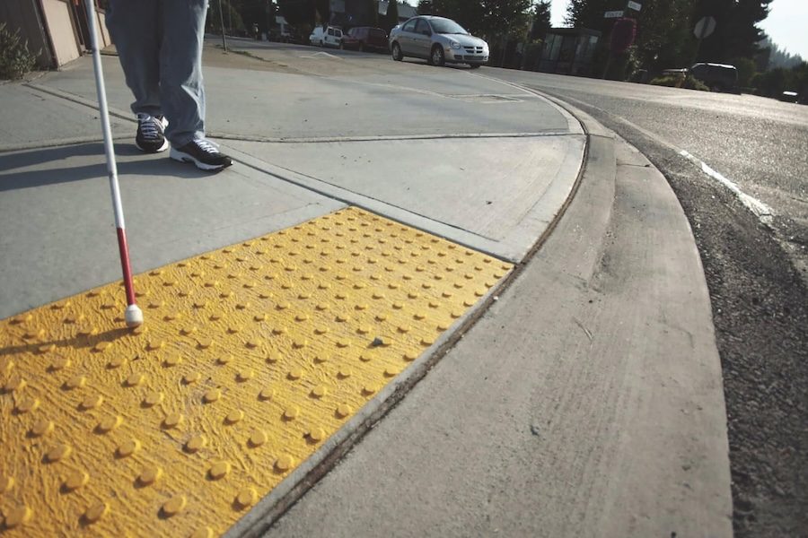 Low perspective of a sidewalk showing yellow tactile bumps, a white cane for a visually-impaired user. You can see a person's legs and black sneakers holding the cane.