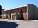 The exterior of a church, photographed in daytime. The building is primarily stone and brick, with a white roof. In front of the church, a paved road is visible. Above the church, the sky is clear and blue.