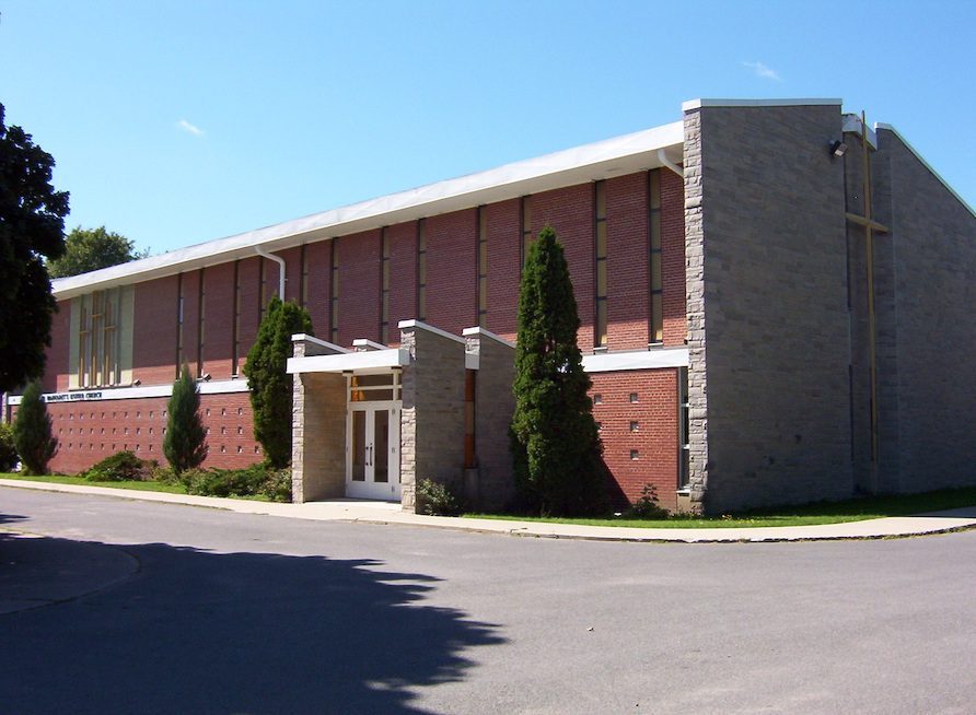 The exterior of a church, photographed in daytime. The building is primarily stone and brick, with a white roof. In front of the church, a paved road is visible. Above the church, the sky is clear and blue.