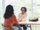 Two women sit across from each other at a table, holding each other's hands.