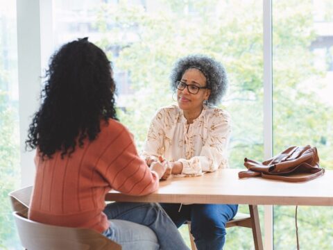 Two women sit across from each other at a table, holding each other's hands.