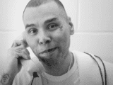 A close-up image of a man holding a landline phone to his ear, smiling and looking at the camera. The photograph is in black and white.