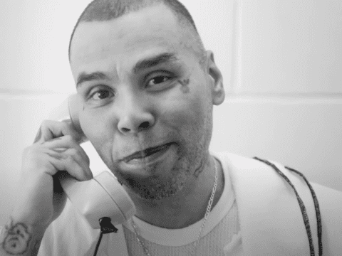 A close-up image of a man holding a landline phone to his ear, smiling and looking at the camera. The photograph is in black and white.