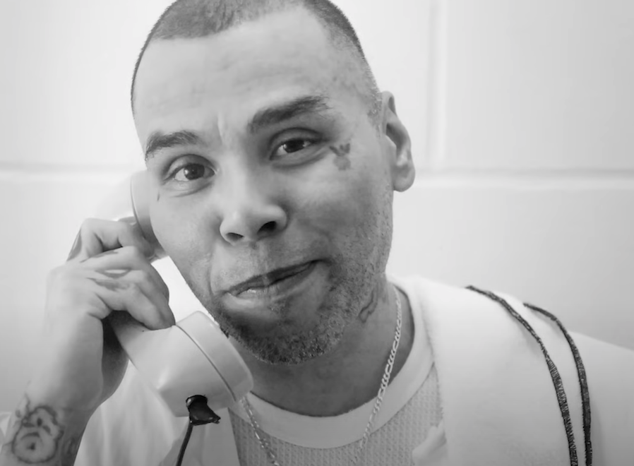 A close-up image of a man holding a landline phone to his ear, smiling and looking at the camera. The photograph is in black and white.