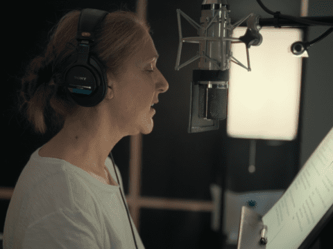 Side profile of Celine Dion, who is facing a microphone and wearing headphones. In front of her seems to be a music stand with sheet music.