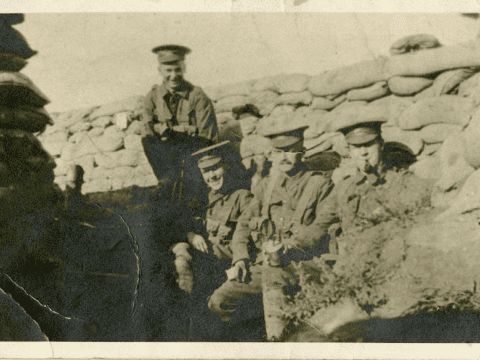 Black and white old photo image of four soldiers sitting in the barracks. Three are seated in front and one behind. They are all in uniform wearing hats. One of them is smoking a pipe. The two on the left are smiling.