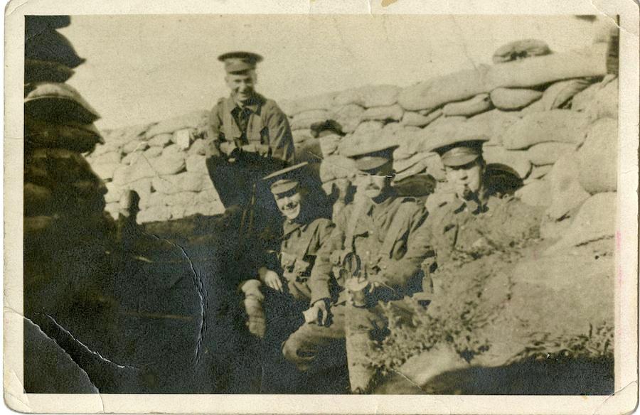 Black and white old photo image of four soldiers sitting in the barracks. Three are seated in front and one behind. They are all in uniform wearing hats. One of them is smoking a pipe. The two on the left are smiling.