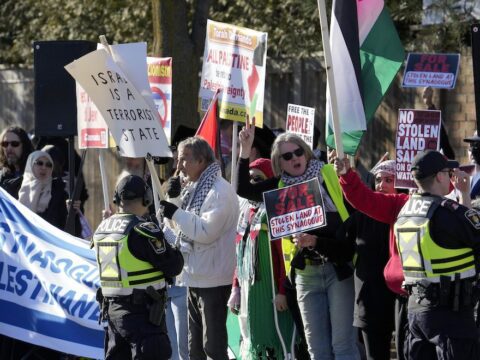 Protesters hold signs facing their camera. Two police officers in vests face stand in front of the protesters, facing away from the camera.
