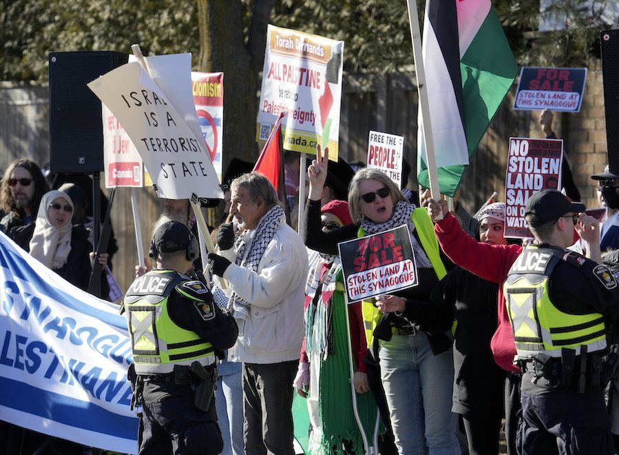 Protesters hold signs facing their camera. Two police officers in vests face stand in front of the protesters, facing away from the camera.