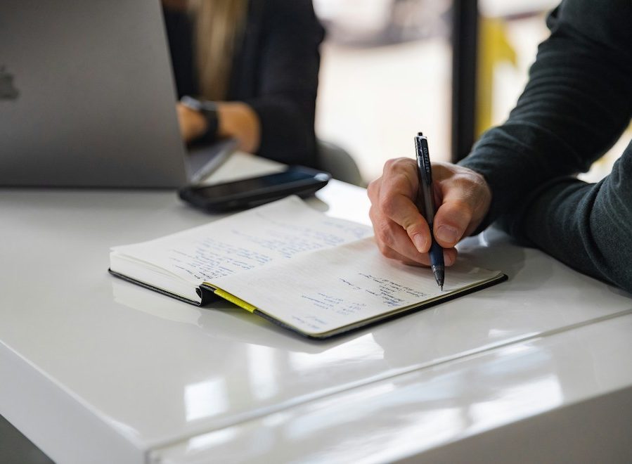 A hand with a pen writing in a notebook on a white desk.