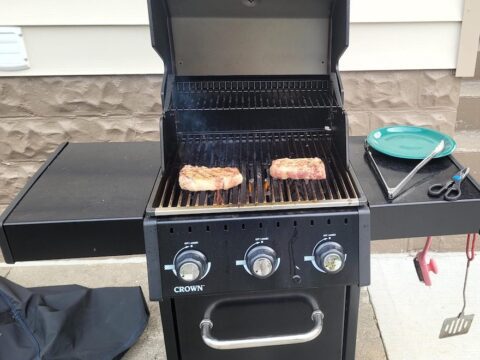 Two steaks cook on top of an open barbecue. A plate, tongs and scissors rest on the right side of the grill.