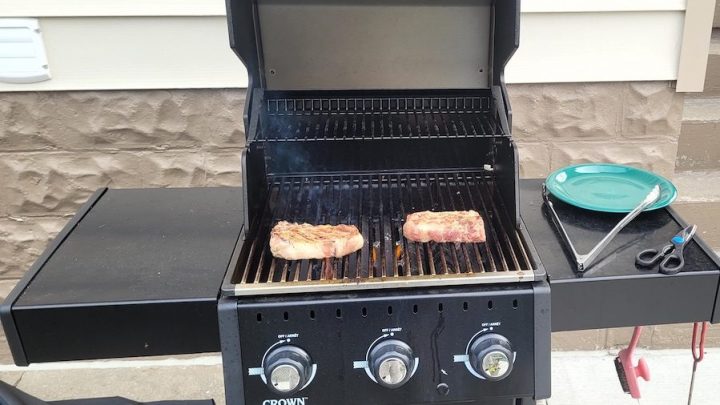 Two steaks cook on top of an open barbecue. A plate, tongs and scissors rest on the right side of the grill.
