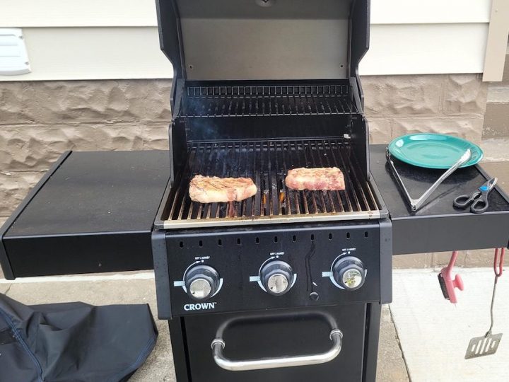 Two steaks cook on top of an open barbecue. A plate, tongs and scissors rest on the right side of the grill.