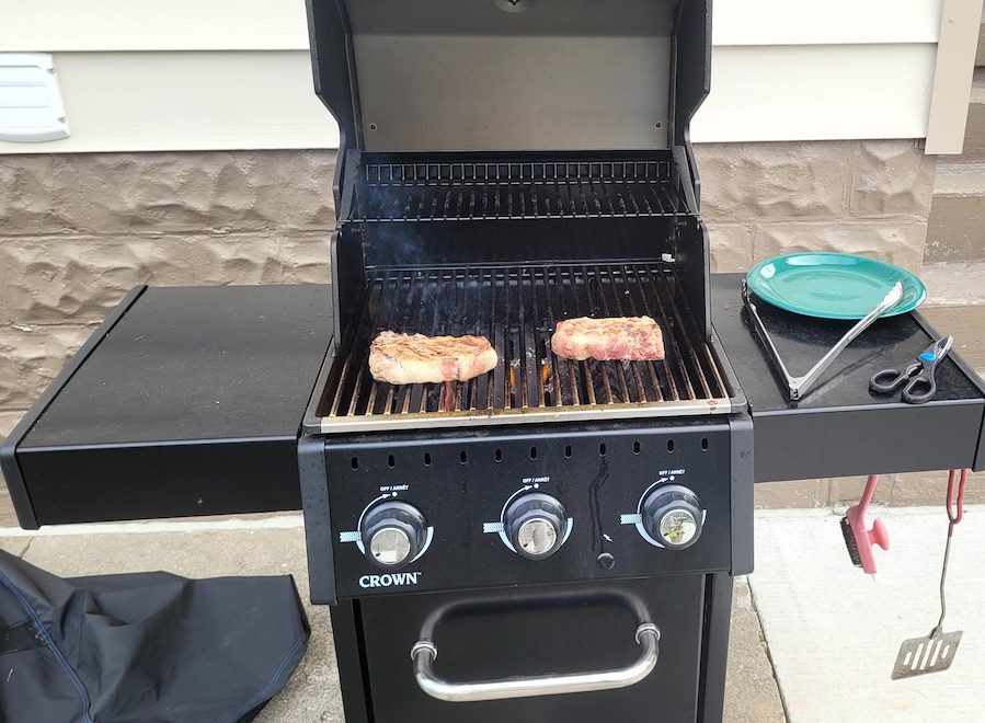 Two steaks cook on top of an open barbecue. A plate, tongs and scissors rest on the right side of the grill.