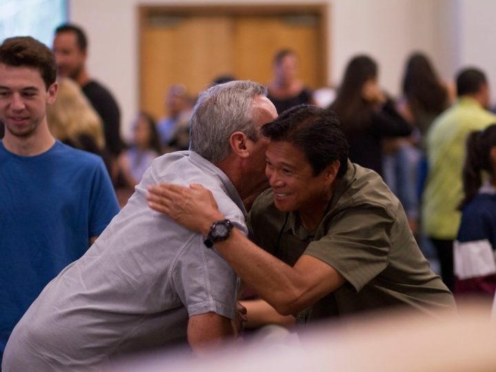 Two men are shaking hands and hugging each other in a room. The other people are in the background and blurred out. One man is East Asian with black hair and tan skin. He is in an olive-coloured shirt and wearing a black watch on his left hand. The other man is a white man whose face is blocked by the Asian man's face. He has white hair and is wearing a grey shirt.
