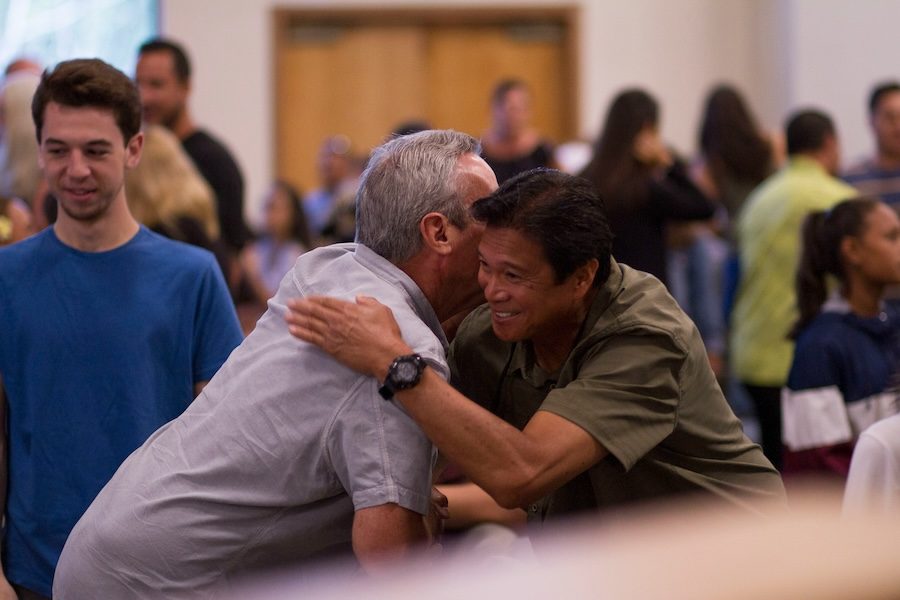 Two men are shaking hands and hugging each other in a room. The other people are in the background and blurred out. One man is East Asian with black hair and tan skin. He is in an olive-coloured shirt and wearing a black watch on his left hand. The other man is a white man whose face is blocked by the Asian man's face. He has white hair and is wearing a grey shirt.