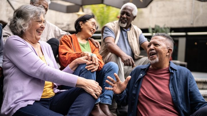 A group of seniors from different backgrounds sit together laughing. There is a Black man with a beard in an orange shirt laughing. Next to him is another Black man with white hair and white beard in a grey shirt and beige vest smiling. Next to him is an Asian woman with black hair, glasses, orange shirt and green t-shirt laughing. Beside her is a white woman with grey hair in a lavender shirt laughing.