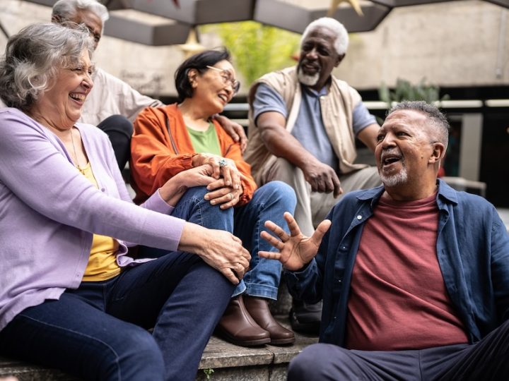 A group of seniors from different backgrounds sit together laughing. There is a Black man with a beard in an orange shirt laughing. Next to him is another Black man with white hair and white beard in a grey shirt and beige vest smiling. Next to him is an Asian woman with black hair, glasses, orange shirt and green t-shirt laughing. Beside her is a white woman with grey hair in a lavender shirt laughing.