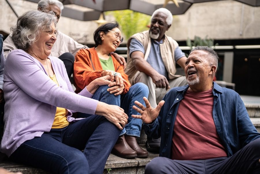 A group of seniors from different backgrounds sit together laughing. There is a Black man with a beard in an orange shirt laughing. Next to him is another Black man with white hair and white beard in a grey shirt and beige vest smiling. Next to him is an Asian woman with black hair, glasses, orange shirt and green t-shirt laughing. Beside her is a white woman with grey hair in a lavender shirt laughing.