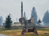 In front, a sign that reads "St. Mary & St. George" fixed into the yellow grass. Behind it, the remains of the church. The air looks smoky.