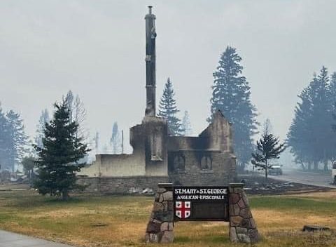 In front, a sign that reads "St. Mary & St. George" fixed into the yellow grass. Behind it, the remains of the church. The air looks smoky.