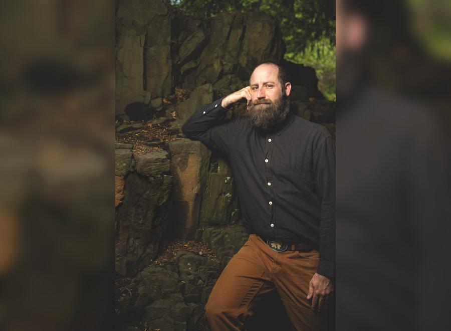 A middle-aged, balding white man poses with his head on his right hand. He is wearing a black button up shirt with brown pants.