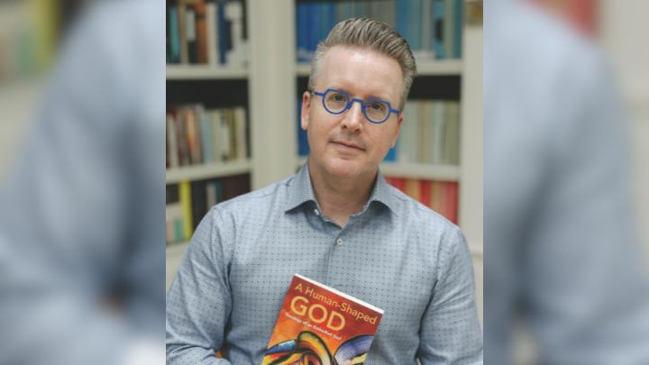 A white man brown and grey hair wearing purple-framed glasses in a blue and brown button-up shirt holds a copy of a book. The title says, "A Human-Shaped God: Theology of an Embodied God."