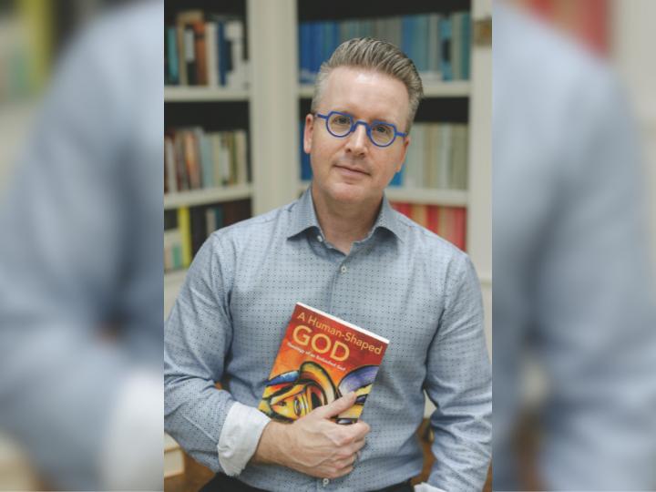 A white man brown and grey hair wearing purple-framed glasses in a blue and brown button-up shirt holds a copy of a book. The title says, "A Human-Shaped God: Theology of an Embodied God."