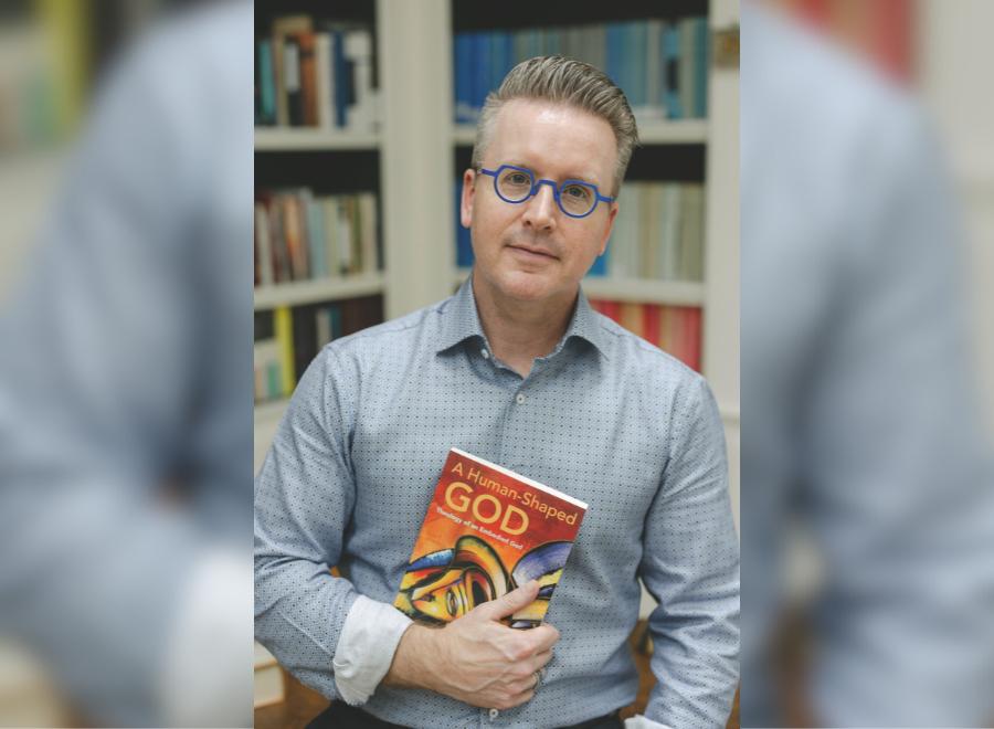 A white man brown and grey hair wearing purple-framed glasses in a blue and brown button-up shirt holds a copy of a book. The title says, "A Human-Shaped God: Theology of an Embodied God."