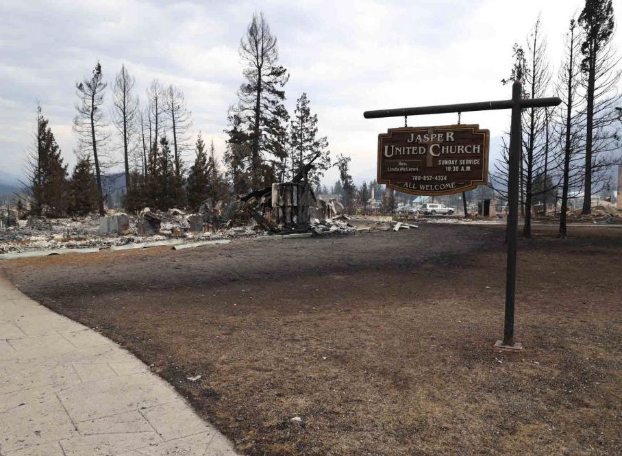 A sign reading "Jasper United Church" sits at the foreground of the image. Behind it, there is debris from the fire.