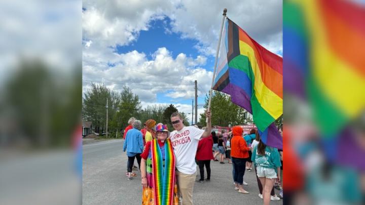 A white woman dressed in a rainbow dress and hat poses next to a young white man in a white shirt and khaki pants. The man is holding a progressive Pride flag and they are at a march.