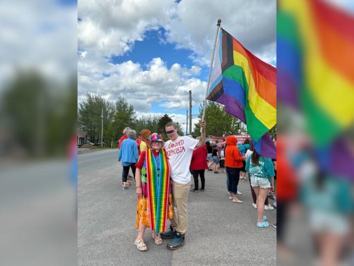 A white woman dressed in a rainbow dress and hat poses next to a young white man in a white shirt and khaki pants. The man is holding a progressive Pride flag and they are at a march.