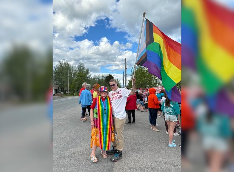 A white woman dressed in a rainbow dress and hat poses next to a young white man in a white shirt and khaki pants. The man is holding a progressive Pride flag and they are at a march.