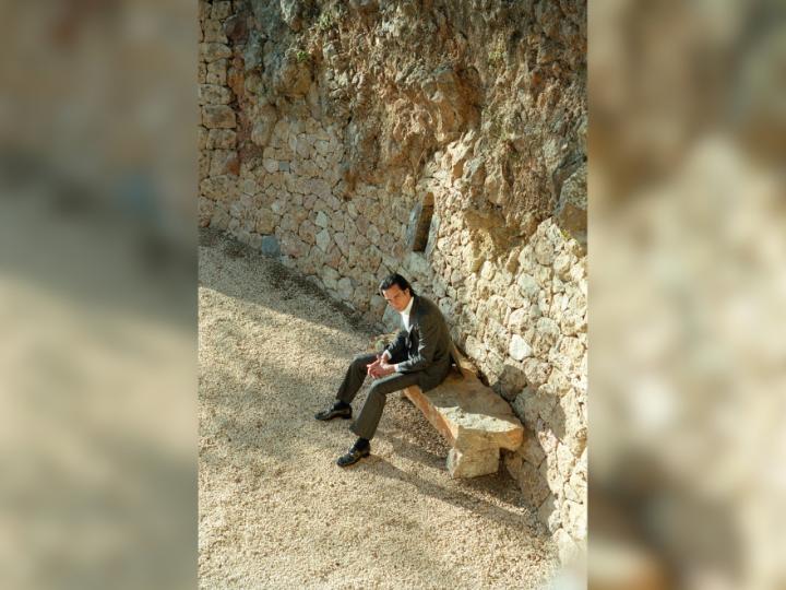 Image of Nick Cave, a white middle-aged man with dark brown hair. He is wearing a black suit with a white button up shirt and sitting on a bench with a rocky background.