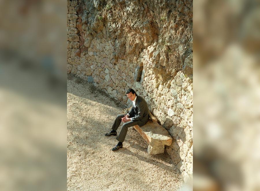 Image of Nick Cave, a white middle-aged man with dark brown hair. He is wearing a black suit with a white button up shirt and sitting on a bench with a rocky background.