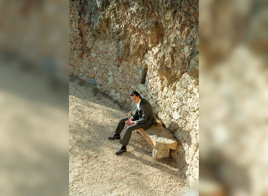 Image of Nick Cave, a white middle-aged man with dark brown hair. He is wearing a black suit with a white button up shirt and sitting on a bench with a rocky background.