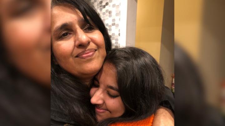 A young brown skin woman in an orange sweater hugs an older brown skin woman with dark brown hair.