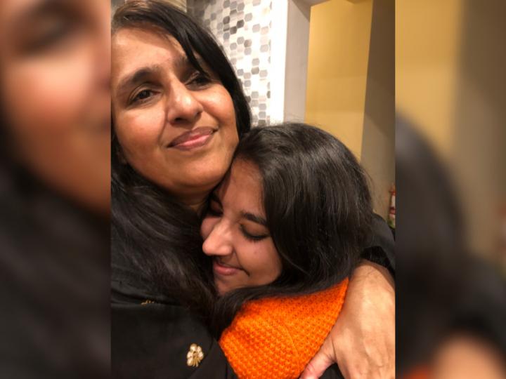 A young brown skin woman in an orange sweater hugs an older brown skin woman with dark brown hair.