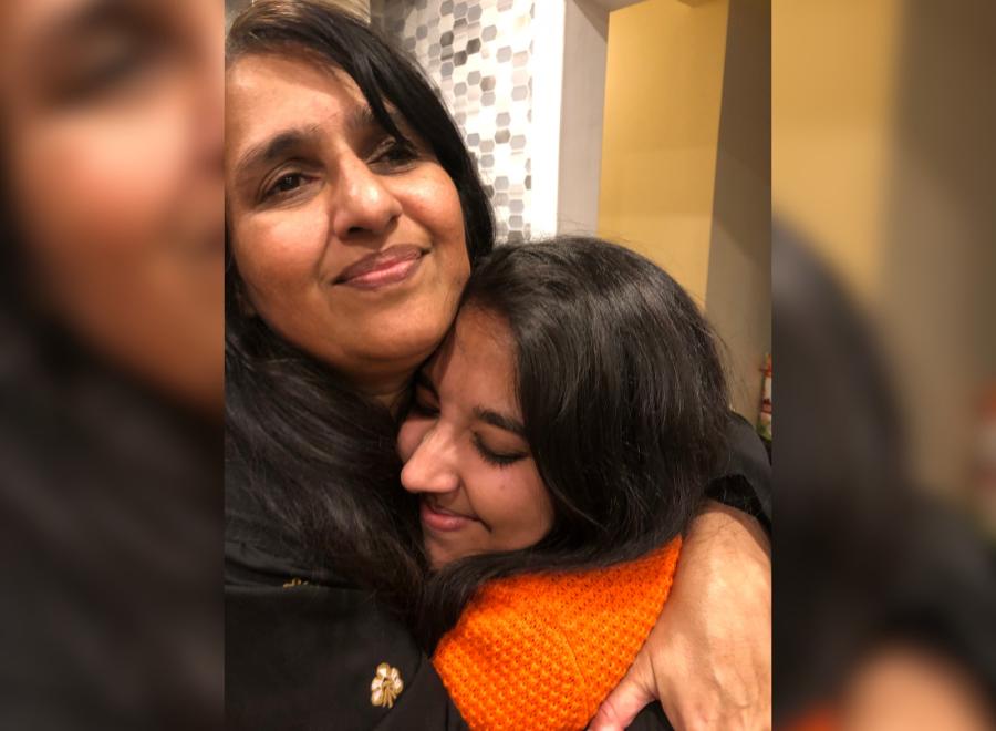 A young brown skin woman in an orange sweater hugs an older brown skin woman with dark brown hair.