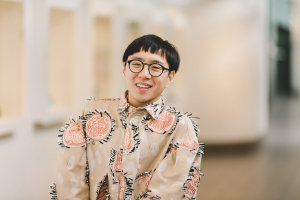 An East Asian man with black hair in a bowl cut smiles. He is wearing round, black-framed glasses and a brown and beige button-up shirt. His shirt has pink circles with black and white fringes around the circles and the buttons. 