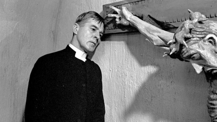 Black and white still from the film "Winter Light". It features a priest staring forlornly next to a crucifixion of Christ.